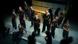 Group of individuals wearing Manchester City 2023 black and yellow kit with Etihad Airways sponsor, standing and sitting indoors.
