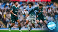 Three soccer players from different teams compete for the ball during a match. The player in blue is wearing a poppy badge on his shirt.