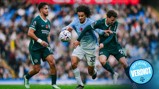 Three soccer players from different teams compete for the ball during a match. The player in blue is wearing a poppy badge on his shirt.