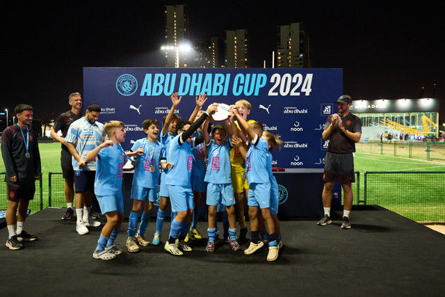 A youth football team celebrating their win at the Abu Dhabi Cup 2024, holding up the trophy on a stage with a large event banner behind them. The team is dressed in blue uniforms.