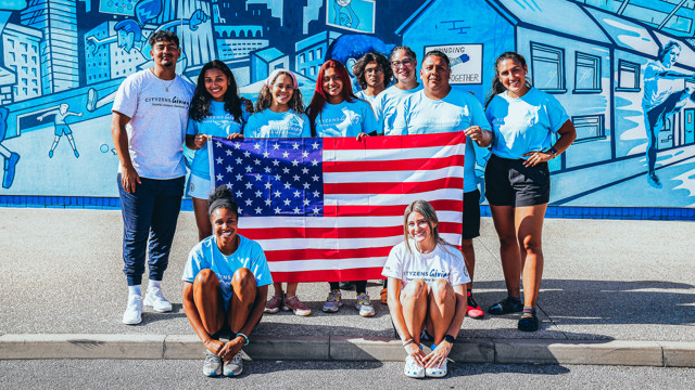 A group of young individuals holding the United States flag in front of a blue mural. They wear T-shirts labeled 'Cityzens Giving Young Leaders Summit'.