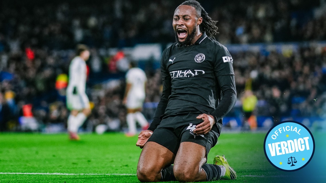 A football player in a black Manchester City kit celebrates a goal by kneeling on the field. The player has long hair, and the game setting includes blurred spectators and another player in the background.