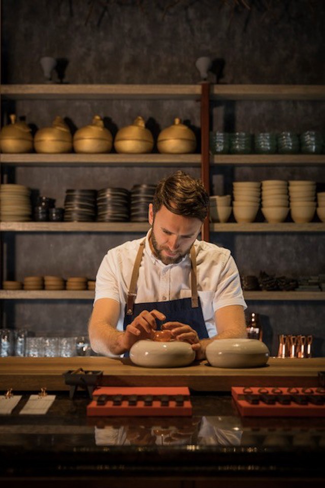 A chef is working behind a counter in a restaurant kitchen. Shelves in the background display various pots and bowls.