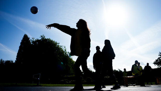 Silhouette of a person throwing a ball outdoors in a park with several people in the background under a bright sun.