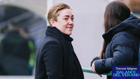 Two individuals have a conversation outdoors during winter. One person wears a black coat with short hair, the other wears a dark winter jacket with long hair. The setting appears to be near sports facilities.