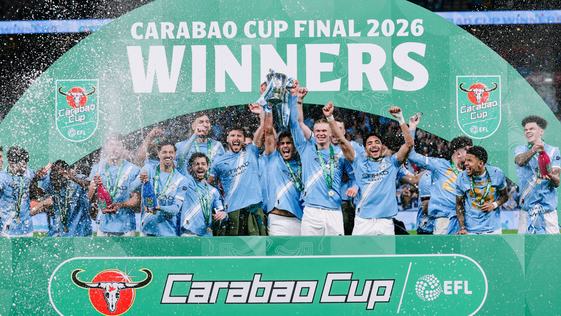 A football team in light blue jerseys celebrates winning the Carabao Cup Final 2026, holding the trophy and spraying champagne.