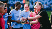 Manchester City women's football team huddling with a coach on the pitch, wearing light blue jerseys.
