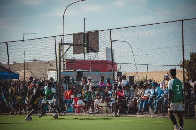 A community soccer match with players in green and black uniforms, surrounded by spectators seated behind a chain-link fence.