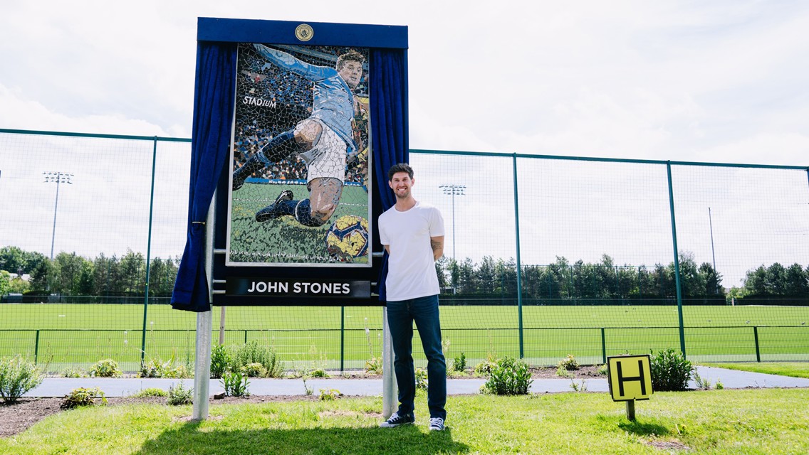 A person stands next to a mosaic portrait of a football player in a sports field, with a sign labeled 'John Stones.'