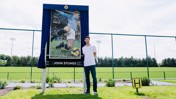 A person stands next to a mosaic portrait of a football player in a sports field, with a sign labeled 'John Stones.'