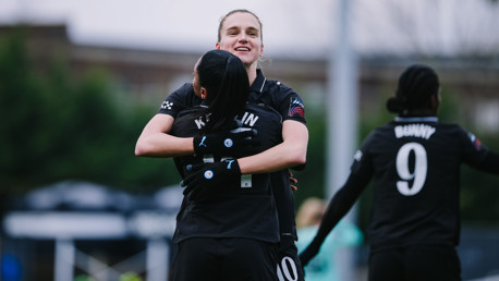 Two female football players embracing on the field, celebrating a moment in what appears to be a Manchester City Women's team match. One player's back shows the name 'Bunny' and number '9'.