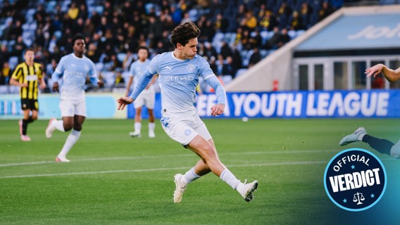 A Manchester City player in action during a soccer match, dressed in the team's light blue kit, about to kick the ball. Other players from Manchester City and the opposing team are visible in the background.