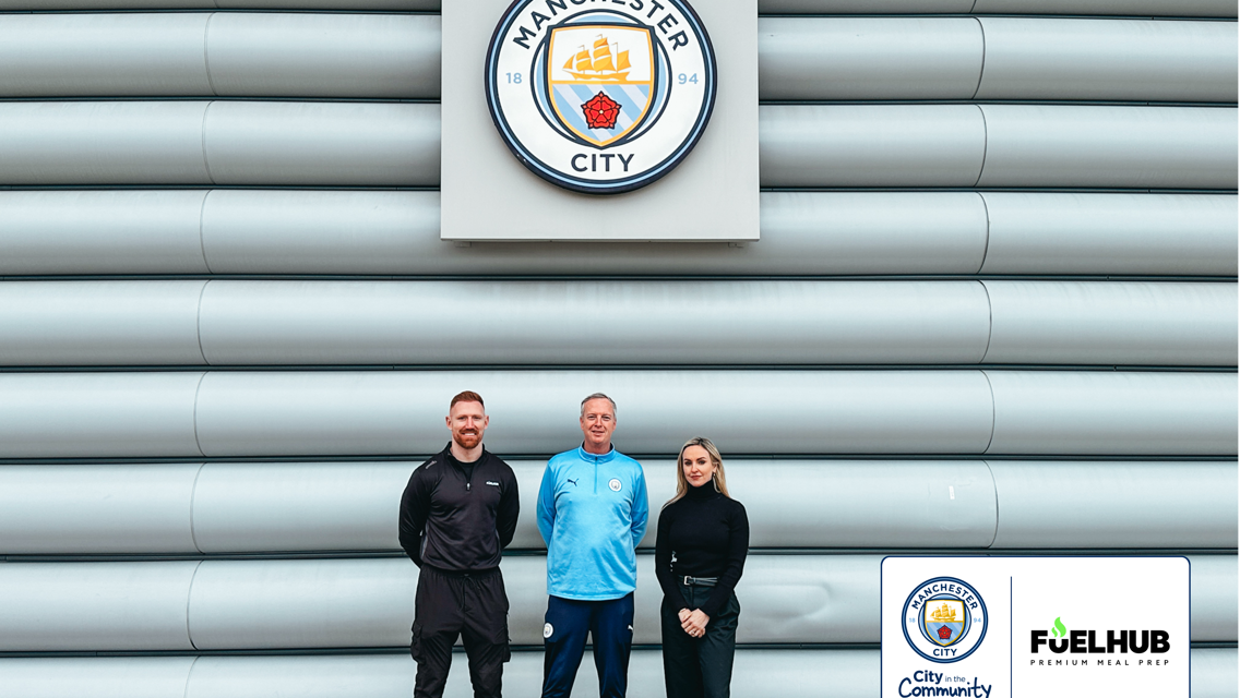 Three individuals stand in front of the Manchester City logo on a wall, next to a partnership announcement with FuelHub for City in the Community.