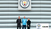 Three individuals stand in front of the Manchester City logo on a wall, next to a partnership announcement with FuelHub for City in the Community.