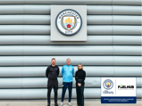 Three individuals stand in front of the Manchester City logo on a wall, next to a partnership announcement with FuelHub for City in the Community.