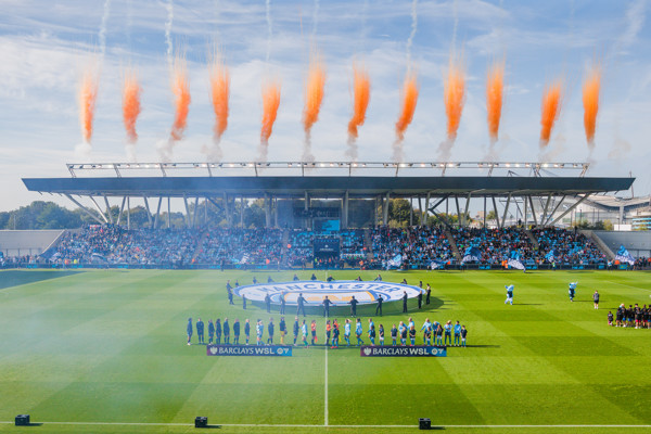 A soccer match at Manchester City's Academy Stadium with teams lined up and pre-match fireworks displaying orange smoke above the stadium.