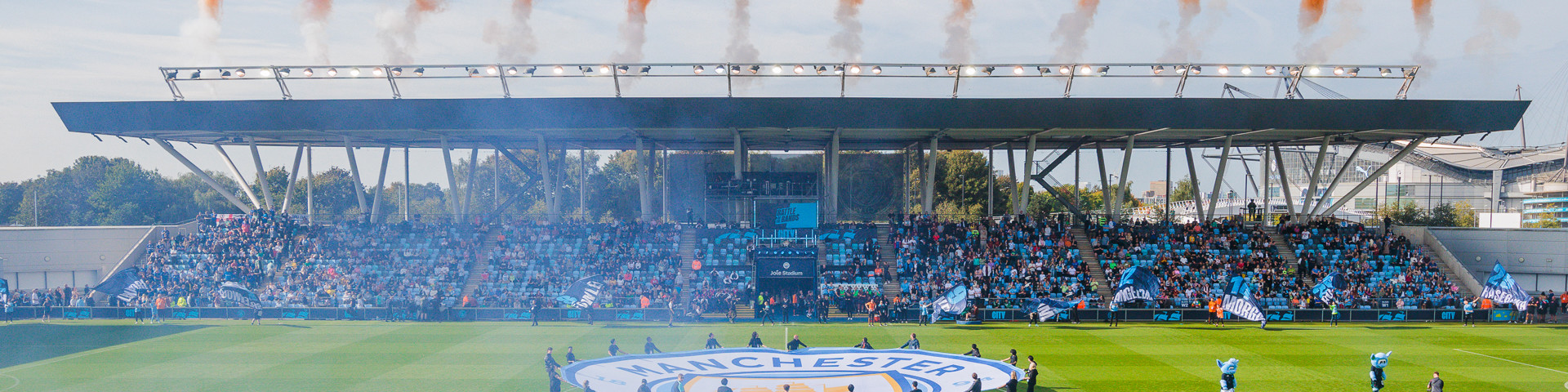 A soccer match at Manchester City's Academy Stadium with teams lined up and pre-match fireworks displaying orange smoke above the stadium.