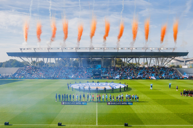 A soccer match at Manchester City's Academy Stadium with teams lined up and pre-match fireworks displaying orange smoke above the stadium.