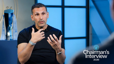 A person giving an interview, wearing a black t-shirt and silver watch with Manchester City branding. A trophy is visible behind them. Text reads: 'Chairman's Interview 2026'.
