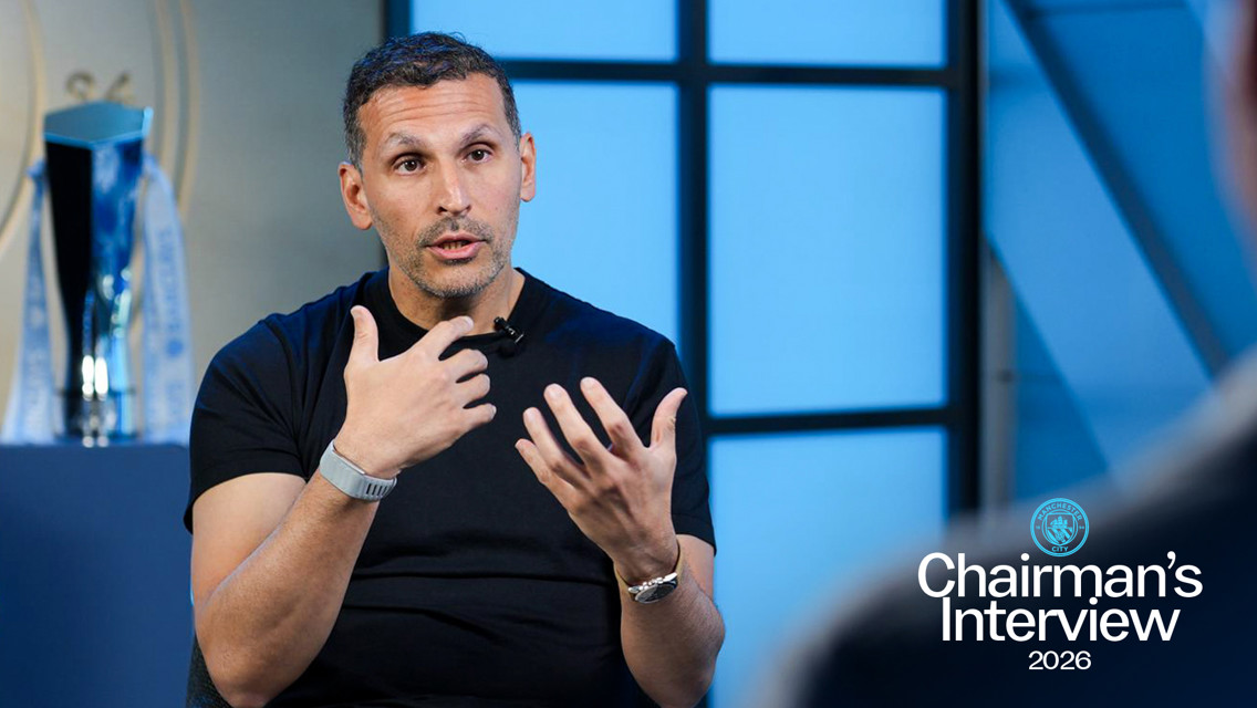 A person giving an interview, wearing a black t-shirt and silver watch with Manchester City branding. A trophy is visible behind them. Text reads: 'Chairman's Interview 2026'.