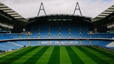 Empty football stadium with blue seats and green pitch, featuring large 'ETIHAD' logo.