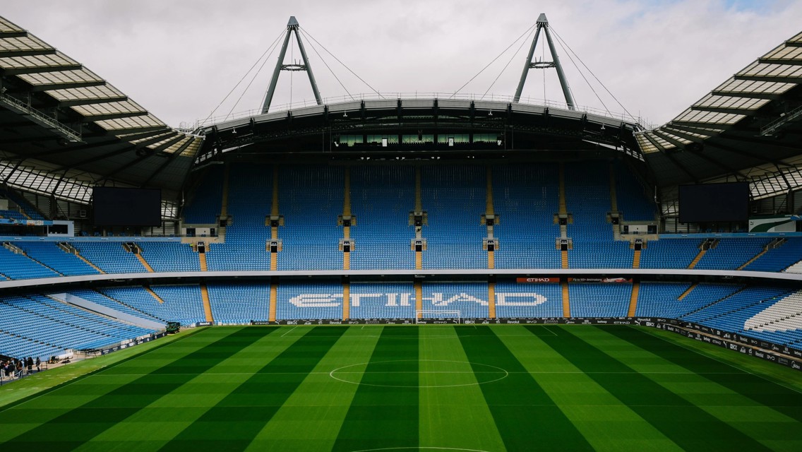 Empty football stadium with blue seats and green pitch, featuring large 'ETIHAD' logo.