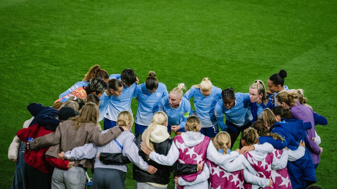 A group of women soccer players in Manchester City jerseys form a team huddle on a soccer field.