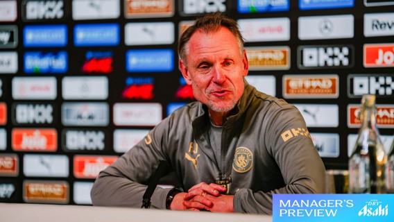 An individual at a press conference wearing a Manchester City jacket, smiling with hands clasped, a microphone in front. Brand logos visible in background.