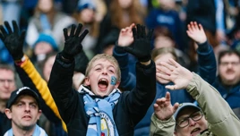 A crowd of soccer fans in a stadium, raising their hands in support. One fan is wearing a Manchester City scarf.