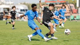 Two youth soccer players in blue and black kits compete for the ball during a match, with blurred faces. Other players and spectators are visible in the background.