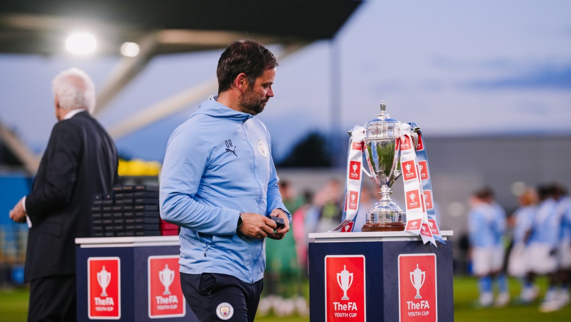 Man in Manchester City attire stands near the FA Youth Cup trophy on a platform with red and white ribbons, while others are blurred in the background.