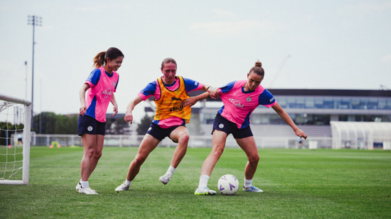 Three female soccer players in pink and blue training uniforms engage in practice on a field. One player is dribbling a ball while others defend.