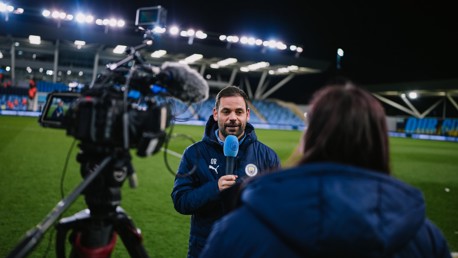 A person in a Manchester City jacket is holding a microphone during a field interview. A cameraman is filming.