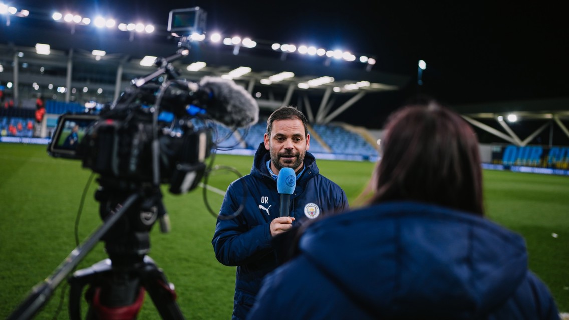 A person in a Manchester City jacket is holding a microphone during a field interview. A cameraman is filming.