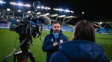 A person in a Manchester City jacket is holding a microphone during a field interview. A cameraman is filming.
