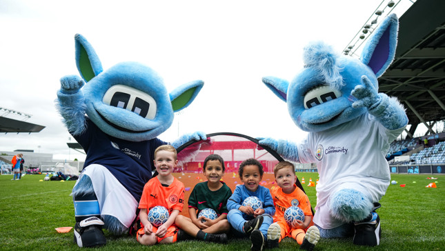Children with footballs pose with Manchester City's mascots, Moonbeam and Moonchester, at an outdoor event hosted by Manchester City at the Etihad Campus.