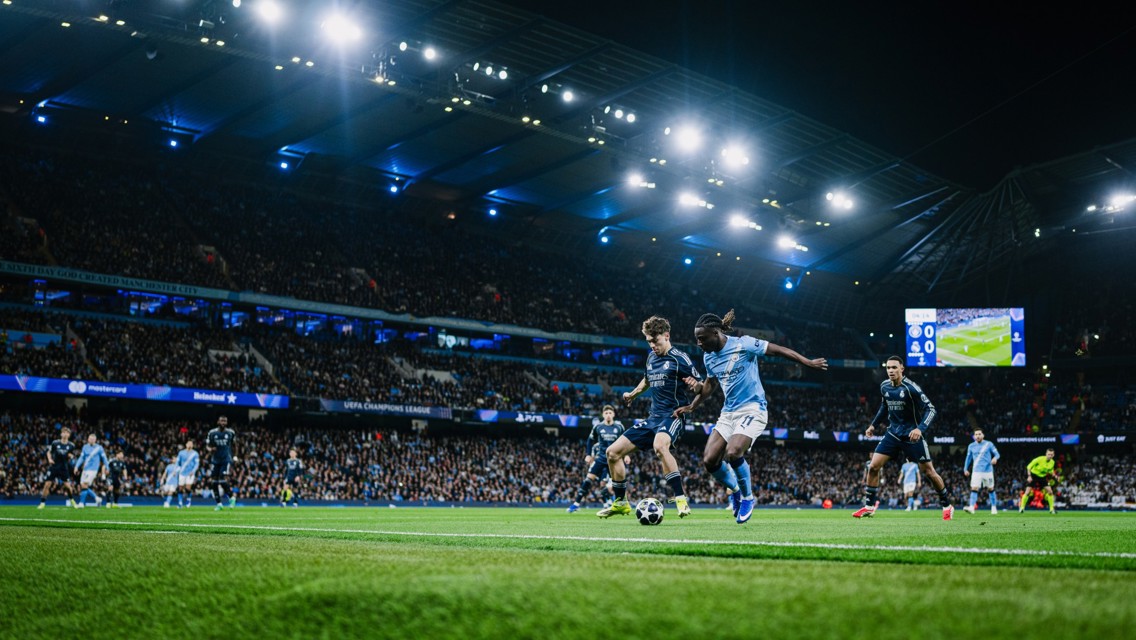 Soccer match between teams in blue and dark jerseys at a stadium, with a UEFA Champions League scoreboard visible.