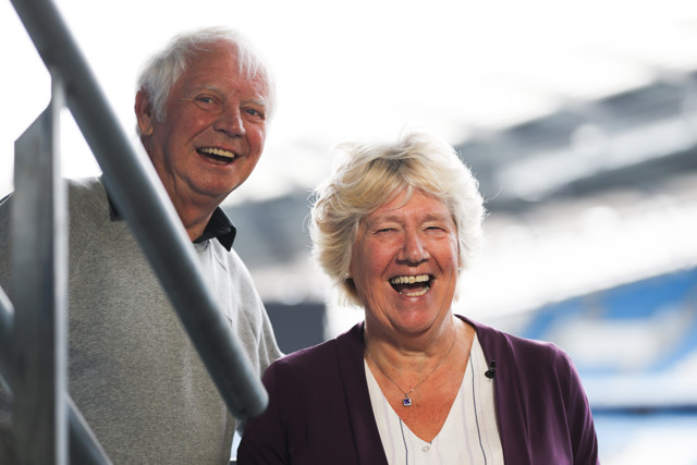 An elderly couple with blurred faces standing in a stadium, with a focus on the woman in the foreground wearing a purple cardigan.