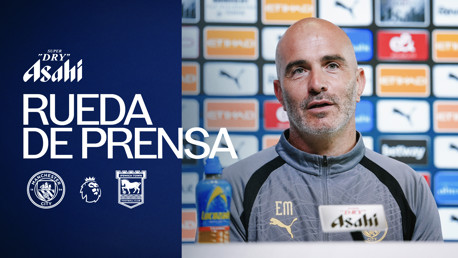A man in a grey Puma tracksuit sits at a press conference table. Logos of Manchester City, Ipswich Town, and the Premier League are displayed. A bottle of Lucozade is in front of him.