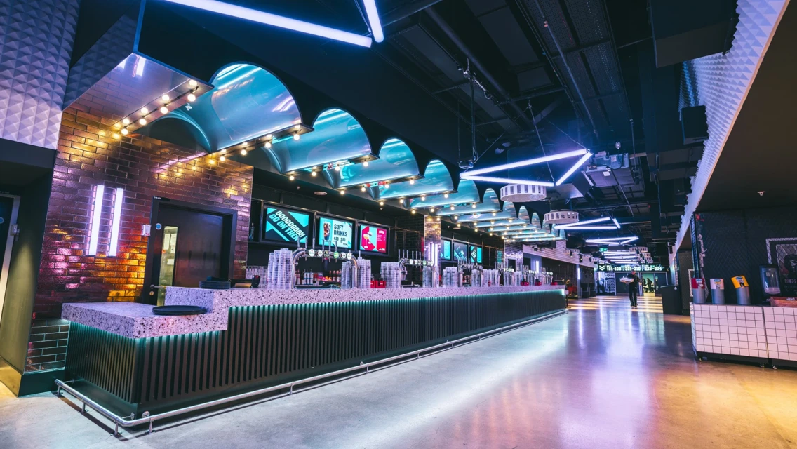 A modern bar setup with neon lights and digital displays. The ambiance is futuristic with glowing elements and a long bar counter filled with stacked cups. The ceiling design and lighting add to the vibrant atmosphere.