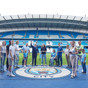 A group of people standing in the Etihad Stadium, home of Manchester City FC, with the Manchester City logo on the ground.