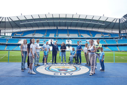 A group of people standing in the Etihad Stadium, home of Manchester City FC, with the Manchester City logo on the ground.