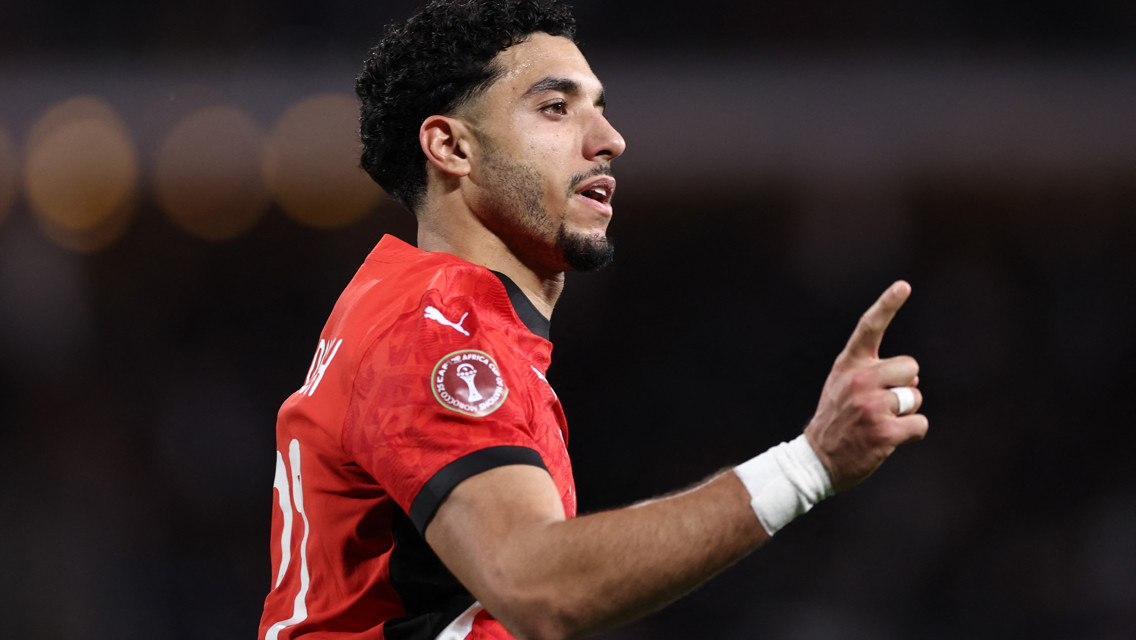 A football player in a red Puma kit pointing upwards, with a CAF Africa Cup of Nations badge visible on the sleeve.
