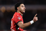 A football player in a red Puma kit pointing upwards, with a CAF Africa Cup of Nations badge visible on the sleeve.