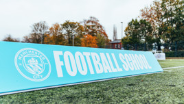 A close-up of a sign at the Manchester City Football School training pitch. The sign features the Manchester City club logo and the text 'FOOTBALL SCHOOL'. Trees and a building are visible in the background.