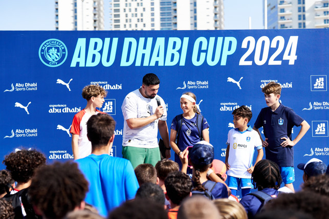 A group of young football players and a presenter at the Abu Dhabi Cup 2024 event, organized by Manchester City and other sponsors, standing in front of a branded backdrop.