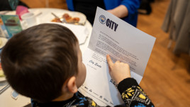 A boy is reading a letter on 'CITY' letterhead at a dining table with food. He is pointing at the text on the letter.