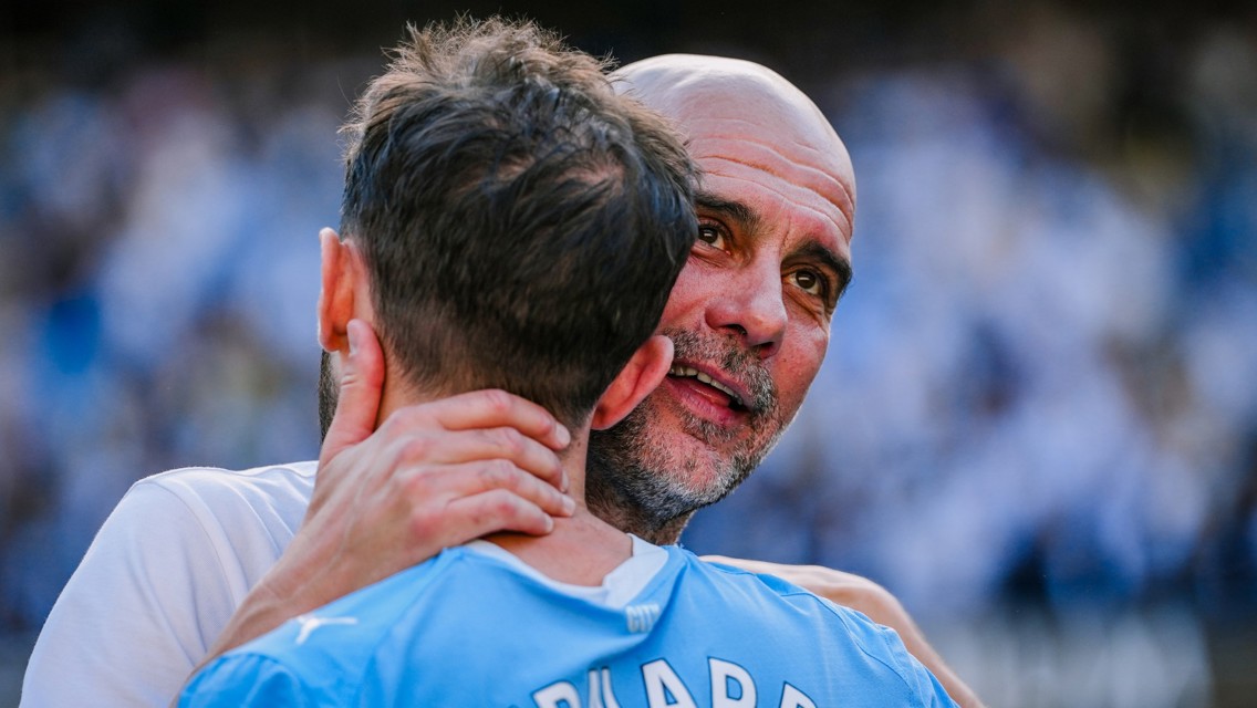 Two Manchester City players embracing on the field, one wearing a blue jersey with 'City' printed on it.
