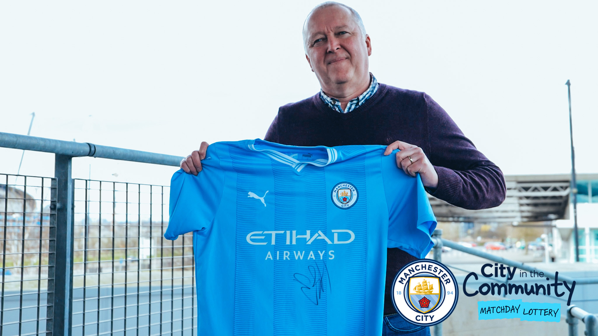 A man holding a signed Manchester City jersey with the City in the Community Matchday Lottery logo.