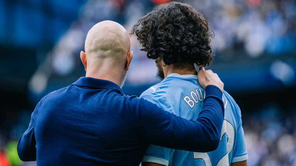 A bald coach places a hand on the shoulder of a curly-haired football player wearing a blue jersey with the name 'BOBB' visible, conveying encouragement.
